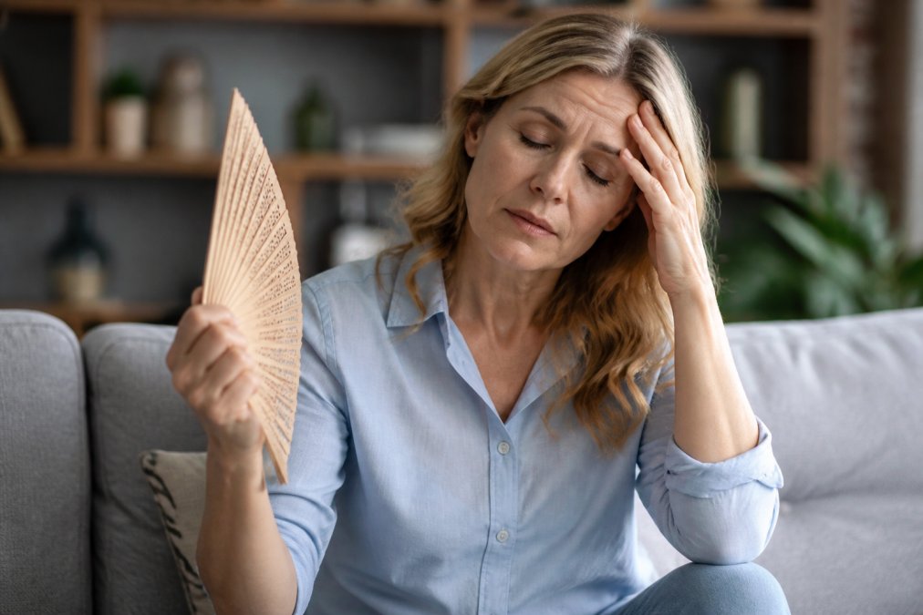 Woman sitting on a gray sofa fanning herself with a wooden hand fan, brow creased in worry.