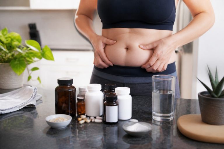 supplements-for-managing-cortisol-and-belly-fat-1787542682 - Thrive Health Solutions Person in a sports bra pinching their belly fat at a kitchen counter with pill bottles nearby and a glass of water