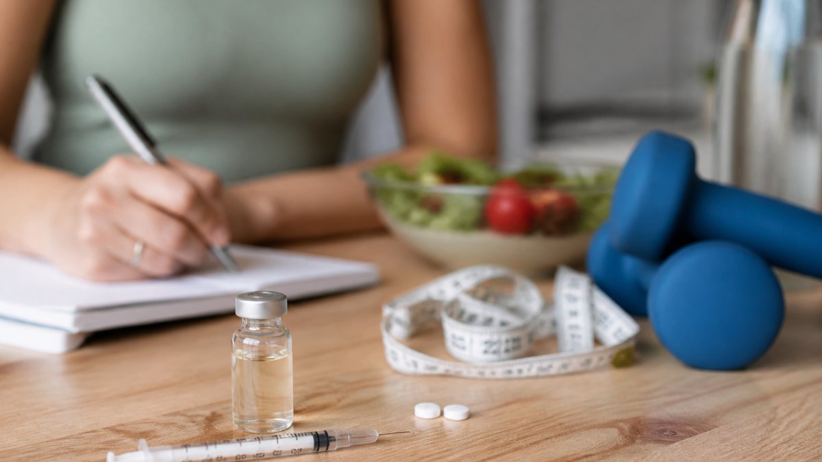 hormone-replacement-therapy-and-weight-management-1790720043-1 - Thrive Health Solutions - Thrive Health Solutions Person writing notes at a table with health items: salad bowl, measuring tape, blue dumbbells, syringe, pills, and a bottle nearby.