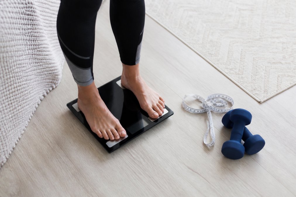 how-long-to-lose-20-lbs-on-ozempic-a-timeline-guide-1789683083-1 - Thrive Health Solutions - Thrive Health Solutions Person standing on a digital bathroom scale barefoot, with a measuring tape and blue dumbbells on the floor nearby