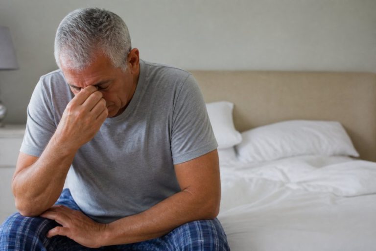 Middle-aged man sitting on the edge of a bed, head in his hand, looking distressed in a bedroom setting.