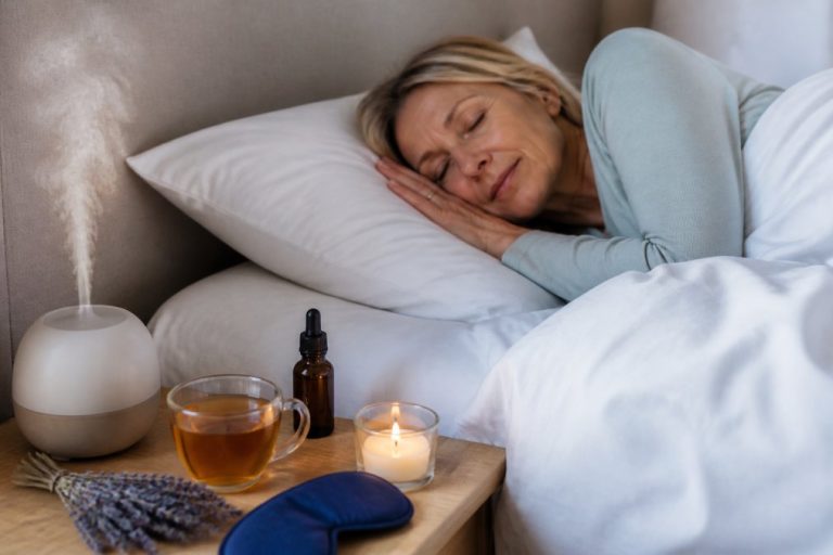 Woman sleeping in bed with a diffuser, cup of tea, lit candle, lavender sprigs, dropper bottle, and eye mask on the bedside table.