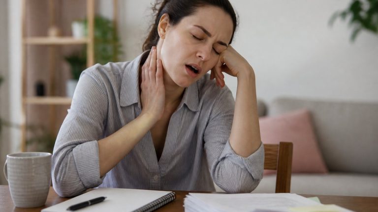 Woman resting her head on her hand and touching her throat, showing throat pain at a desk with papers and a mug nearby