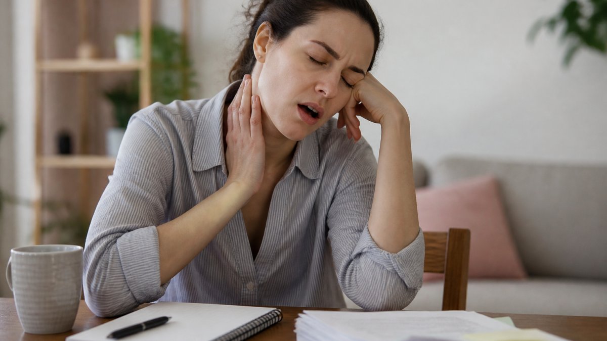 signs-of-thyroid-imbalance-causing-exhaustion-1791844269-1 - Thrive Health Solutions - Thrive Health Solutions Woman resting her head on her hand and touching her throat, showing throat pain at a desk with papers and a mug nearby